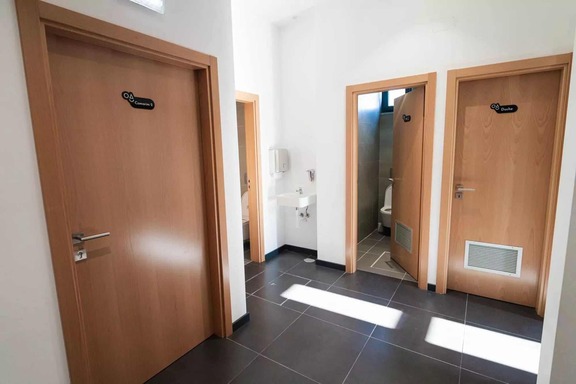 Corredor de acesso aos balneários e camarins com portas de madeira, lavatório e piso em cerâmi...