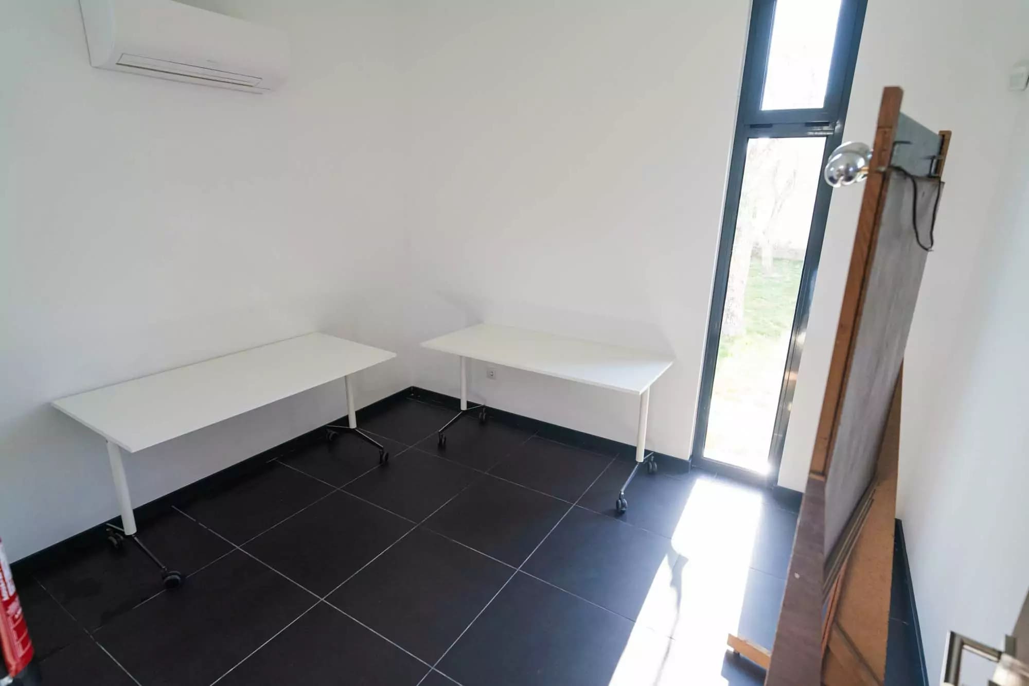 Interior de uma sala de apoio com paredes brancas, duas mesas com rodas, espelho de camarim e jan...